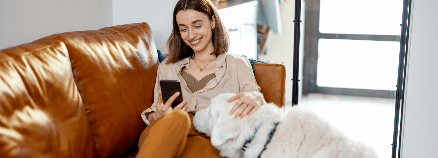 Woman sitting on a brown leather couch using a smartphone, with a white dog beside her.