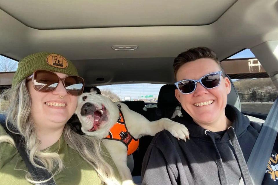 two people in a car with a dog in the back resting it's head on the left person's shoulder