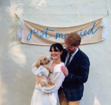 a married couple holding a dog and standing in front of a just married sign hanging on the wall behind them